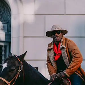 Cowboy in a brown duster and gray hat and sunglasses riding a black horse