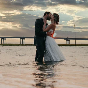 a wedding couple embrace in water with a bridge and sunset in the background