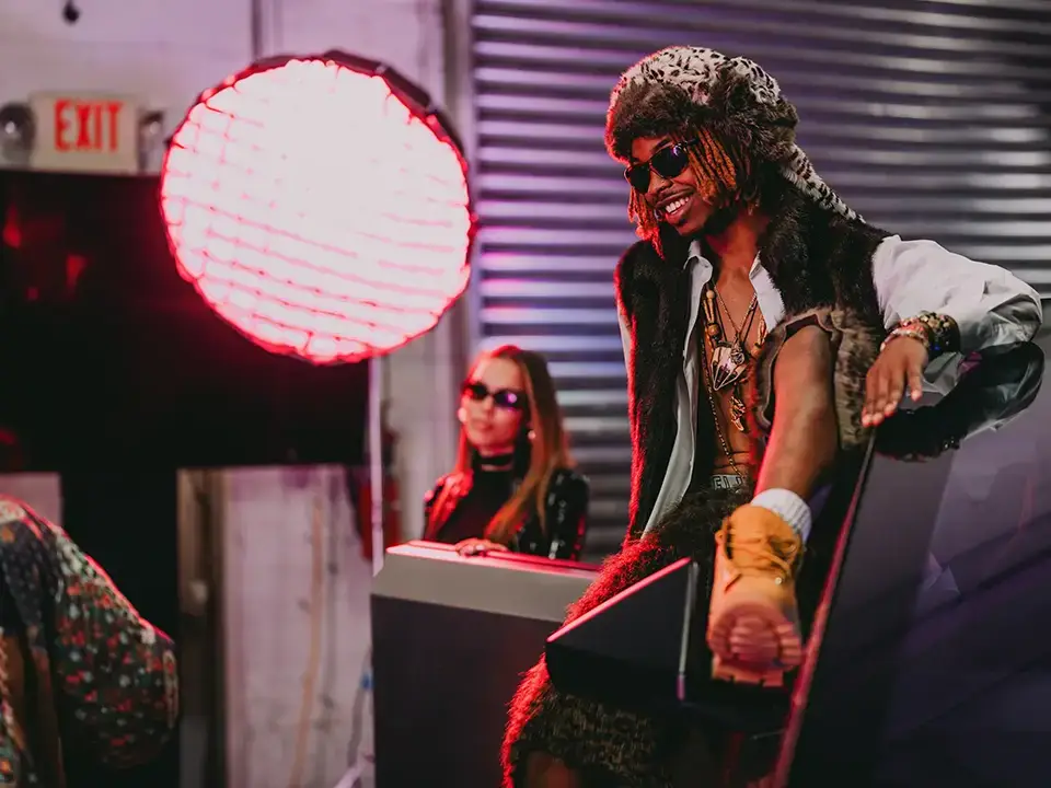 a model with a fuzzy hat and sunglasses poses in front of a studio lighting setup