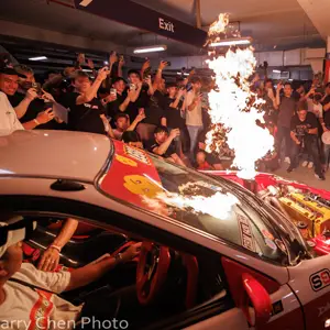 Group gathers cheering around a car with flames coming out of its engine.
