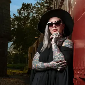 Woman in black dress, hat, and sunglasses, leaning against a red train car